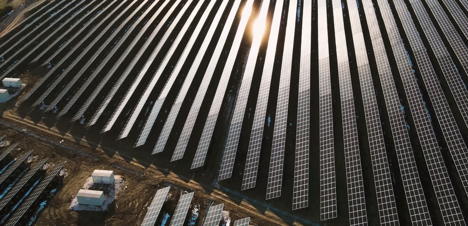 Sobrevoo de uma usina solar ao pôr do sol, fileiras de trackers refletindo a luz